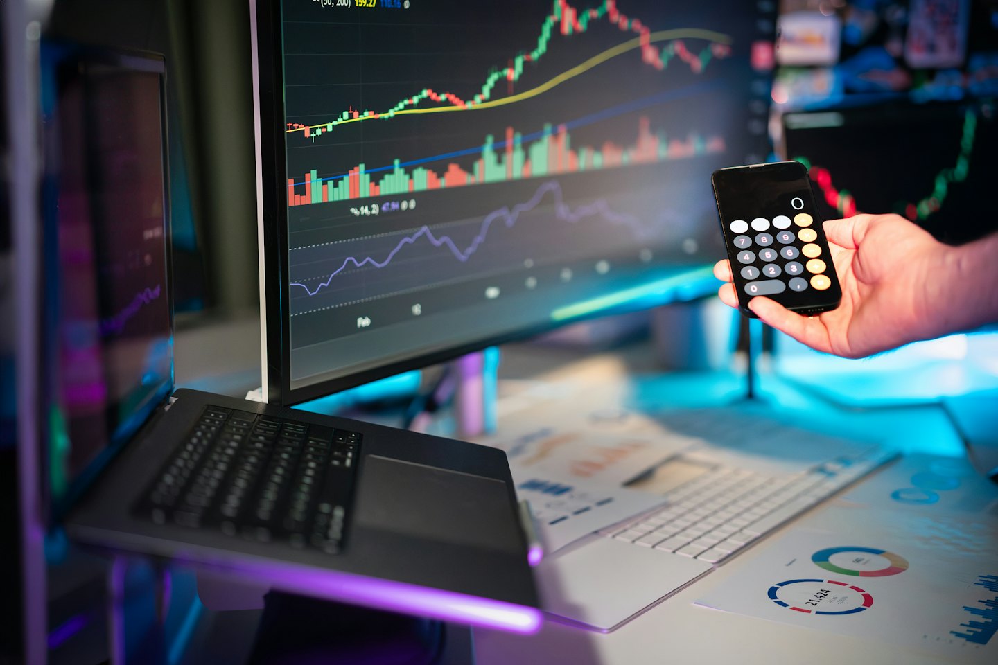 Hand houdt telefoon met rekenmachine vast bij financiële gegevens op monitoren.